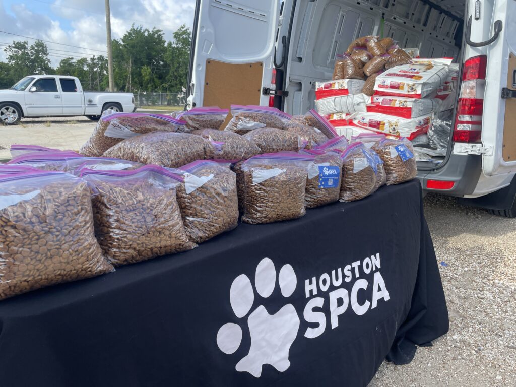 A black tablecloth with the Houston SPCA logo covers a table full of plastic bags of Hill's Pet food. A Houston SPCA sprinter appears in the background with the back open to show many more full bags of food.