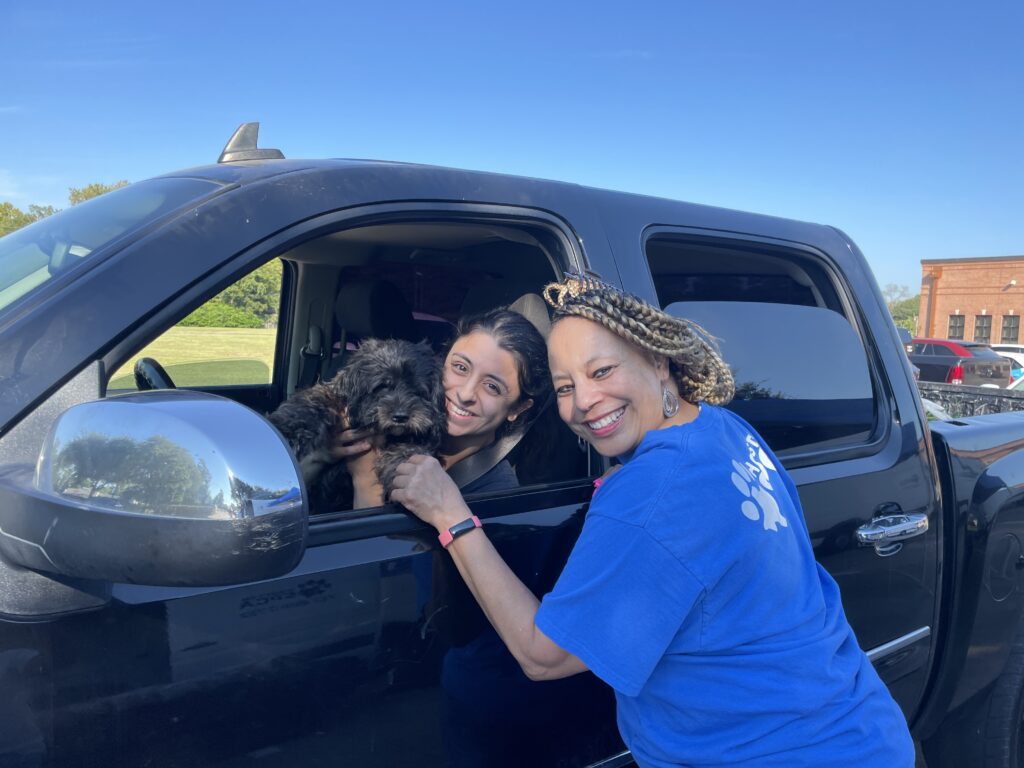 A Houston SPCA volunteer smiles at the truck window of a woman with her dog inside at a community event.