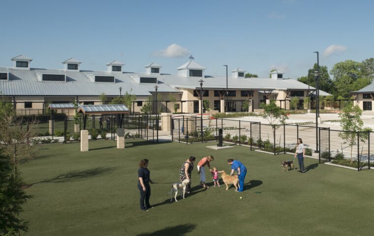 Houston SPCA William & Evelyn Griffin Campus For All Animals aparece en el fondo contra un cielo azul. El edificio tiene un techo gris claro con pequeñas torres con ventanas, una con una veleta con un perro. En primer plano, en un parque verde para perros, se reúnen dos perros y cuatro personas.